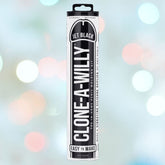 A package labeled "Clone-A-Willy Vibe Kit-Jet Black" by Empire Labs stands against a softly blurred background with pastel circles. The cylindrical packaging is mainly black and white, featuring text that highlights its ease of use.